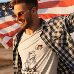 Smiling man wearing sunglasses and a checkered shirt over a gray T-shirt featuring the “Gluttonous Rex” design — a cartoonish T-Rex with a hairstyle resembling Donald Trump, thinking about fried chicken, fries, and soda. The man poses outdoors holding an American flag, symbolizing freedom and satire. Political pop art fashion from the “Political Prehistoric” series by FineArtsLola.